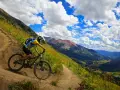 A mountain biker navigates a winding trail, enjoying the breathtaking views of cross country mountain bike trails winding through a lush valley and rugged peaks under a dramatic sky.