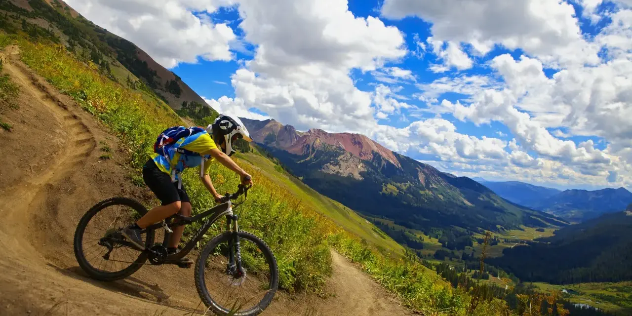 A mountain biker navigates a winding trail, enjoying the breathtaking views of cross country mountain bike trails winding through a lush valley and rugged peaks under a dramatic sky.
