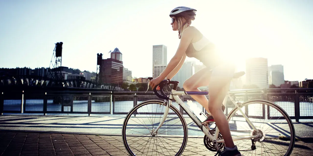 A female cyclist enjoys a sunny ride along a waterfront path with a city skyline in the background.