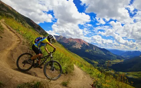 A mountain biker navigates a winding trail, enjoying the breathtaking views of cross country mountain bike trails winding through a lush valley and rugged peaks under a dramatic sky.