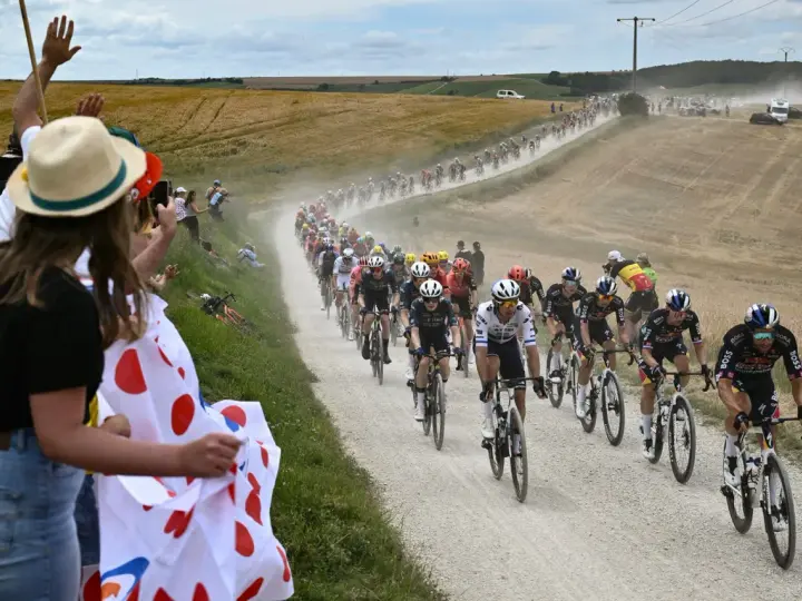 Spectators cheer as a peloton races on a dusty road during the Tour de France watch. Cyclists in colorful jerseys kick up dust in a dramatic scene.