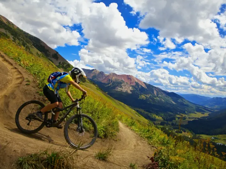 A mountain biker navigates a steep, winding trail, experiencing the best mountain biking in Colorado with stunning valley views and dramatic clouds.