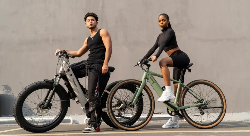A man and woman pose with their electric bikes. This shows what a hybrid bike is good for: versatile riding on various terrains, blending comfort and performance for urban commutes or light trails.