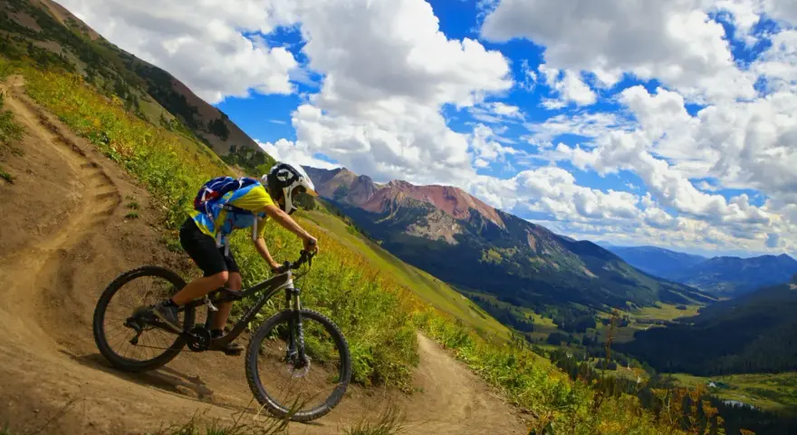 A mountain biker navigates a steep, winding trail, experiencing the best mountain biking in Colorado with stunning valley views and dramatic clouds.